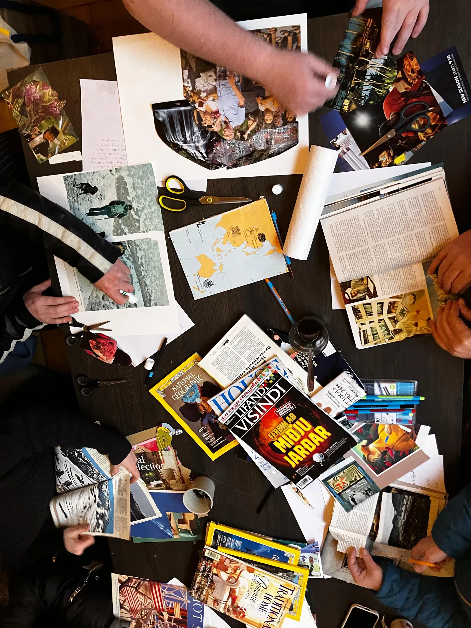 An overhead photograph of the Mental Bytes workshop table, with hands making collages among magazines, coloured paper, scissors and drawing materials